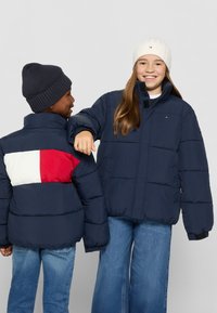 Marina puffjackor med röda och vita detaljer på baksidan, i kombination med blå jeans. Flickan har på sig en vit ribbad mössa, medan pojken har en marinblå.