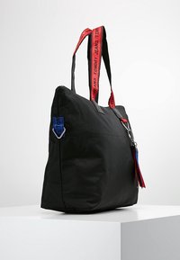 Borsa a mano in nylon nero con manici brandizzati rossi e neri, etichetta accentata blu, chiusura con cerniera e nappine. Forma rettangolare con una texture liscia.