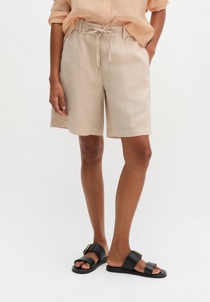 Person trägt beige knieumspielte Shorts mit Kordelzug in der Taille, ein pfirsichfarbenes Hemd und schwarze Sandalen mit offenen Zehen und Schnallen.