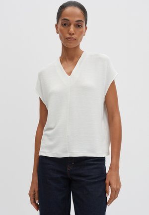 Haut blanc à manches courtes avec un col en V, présentant un tissu texturé et une coupe ample. Associé à un jean bleu foncé. Design simple et épuré.