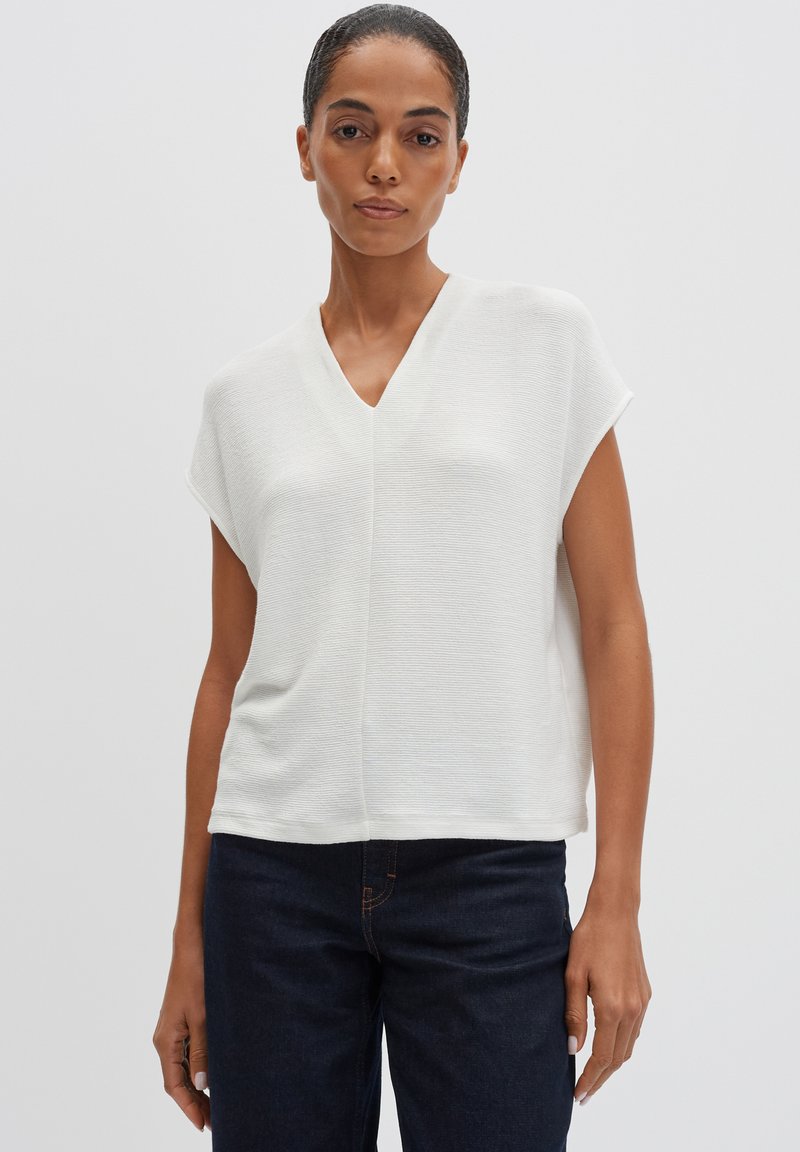 Haut blanc à manches courtes avec un col en V, présentant un tissu texturé et une coupe ample. Associé à un jean bleu foncé. Design simple et épuré.