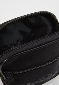 Porte-documents noir à fermeture éclair en tissu durable, présentant une doublure intérieure lisse et un extérieur texturé. Agrémenté d'un détail de logo.