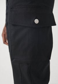 Μαύρο cargo παντελόνι με μεγάλη πρόσθια τσέπη με καπάκι, ασφαλισμένη με ασημένιο κουμπί πίεσης. Το ύφασμα φαίνεται λείο με ελαφριά γυαλάδα.