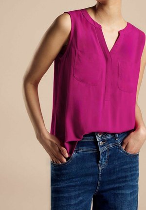 Ærmeløs magenta bluse med forlommer, kombineret med blå denimjeans, hænderne hvilende afslappet i forlommerne.