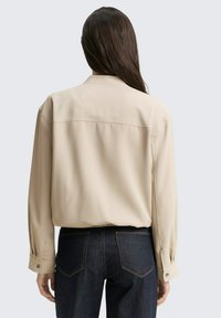 Chemise beige à manches longues avec une coupe décontractée, présentant des poignets à boutons et un détail de couture verticale dans le dos, associée à un jean en denim foncé.