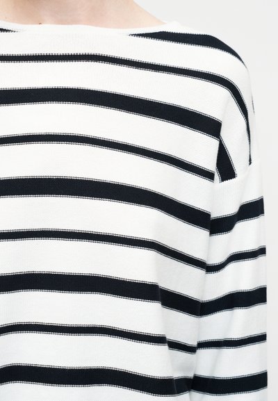 Λευκό πλεκτό πουλόβερ με φαρδιές και στενές οριζόντιες μαύρες ρίγες, εμφανιζόμενο στο πάνω μέρος του κορμού και στον ώμο.