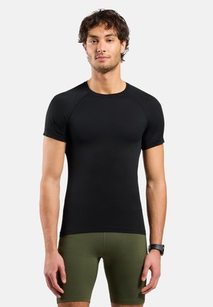 Athletischer Mann steht, trägt ein eng anliegendes schwarzes kurzärmeliges Shirt und olivgrüne Kompressionsshorts, mit einer Smartwatch am linken Handgelenk.