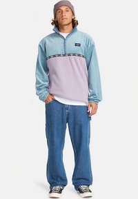 Άντρας ντυμένος με μπλε και μοβ πουλόβερ, φαρδιά μπλε τζιν, ριγέ αθλητικά παπούτσια και μωβ σκούφο, στέκεται με το ένα χέρι στην τσέπη.