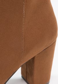 Gros plan d'une botte cheville en daim marron montrant une couture latérale et un talon bloc épais sur fond blanc.