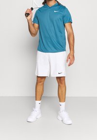 Tyrkysové sportovní tričko Nike se V-výstřihem, kombinované s bílými šortkami s bočními kapsami. Bílé tenisky a černé akcenty Nike.