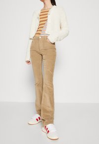 Cardigan flou off-white, haut rayé multicolore, pantalon en velours côtelé beige évasé, et baskets blanches avec des accents rouges.