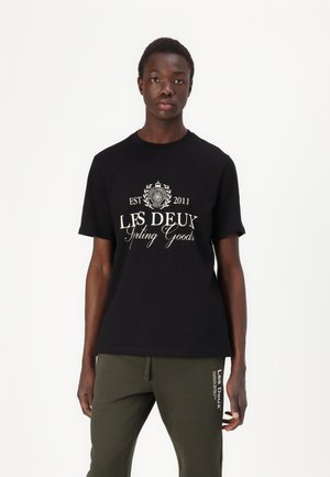 Black cotton t-shirt with a round neck, featuring white text logo "LES DEUX" and a decorative emblem. Worn with olive green sweatpants.