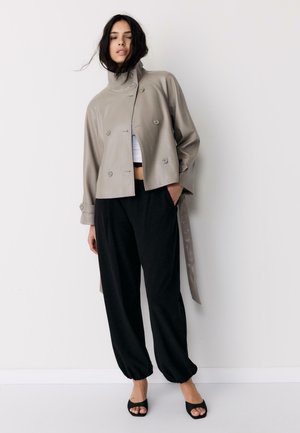 Vrouw met losse zwarte broek, taupekleurige dubbelgeknoopte cropped jas, wit cropped topje en zwarte open tenen hakken tegen een effen lichte achtergrond.