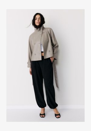 Vrouw met losse zwarte broek, taupekleurige dubbelgeknoopte cropped jas, wit cropped topje en zwarte open tenen hakken tegen een effen lichte achtergrond.