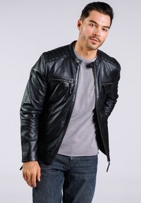 Chaqueta de cuero negra con cuello levantado, dos bolsillos en el pecho con cremallera y un diseño estilizado. Combinada con una camiseta gris de cuello redondo y jeans de mezclilla.