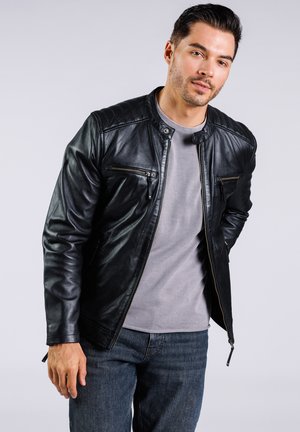 Chaqueta de cuero negra con cuello levantado, dos bolsillos en el pecho con cremallera y un diseño estilizado. Combinada con una camiseta gris de cuello redondo y jeans de mezclilla.