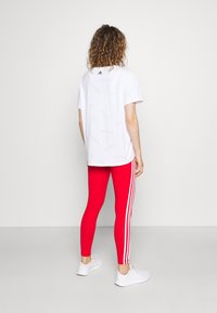 Biała koszulka z delikatnym wzorem logo Adidas, połączona z jasnoczerwonymi legginsami z białymi paskami po bokach oraz białymi trampkami.