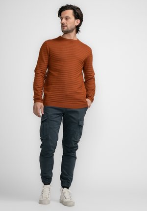 Rust-colored ribbed sweater with round neckline, paired with dark cargo pants featuring multiple pockets and elastic cuffs. White sneakers complete the look.