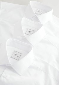 Trzy chrupiące białe koszule z kołnierzykiem zapinanym na guziki, z metkami brzmiącymi "NEXT TAILORING". Gładka tekstura i jednolity design.