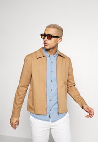 Beige bomulljakke med krage, lagd over en lyseblå denimskjorte. Modellen har på seg mørke solbriller, stylet med hvite bukser.