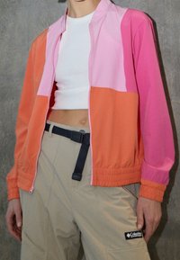 Veste color block en orange, rose et rose clair, fabriquée en matériau léger. Dotée d'une fermeture éclair à l'avant et de poignets élastiques. Associée à un pantalon beige.