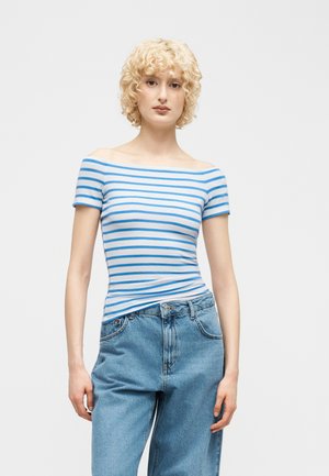 Giovane con capelli biondi corti e ricci, indossa una maglia a righe blu e bianca con spalle scoperte e jeans blu larghi, su sfondo bianco.