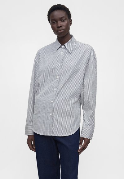 Άτομο που φοράει άνετο λευκό πουκάμισο με μακριά μανίκια, κουμπωτό, με λεπτές μαύρες κάθετες ρίγες και σκούρο μπλε παντελόνι, μπροστά σε μονόχρωμο φόντο.