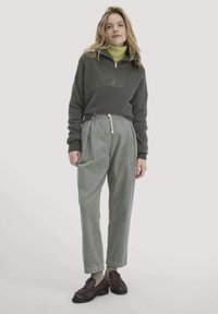 Dunkelgrüner Pullover mit hohem Kragen und Reißverschluss, kombiniert mit hellgrauen gestreiften Hosen und dunkelbraunen Schuhen; die Texturen kontrastieren dezent.