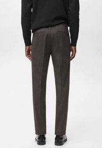 Pantaloni su misura marrone scuro con una texture liscia, caratterizzati da un taglio dritto e due tasche posteriori. Calzature nere completano il look.