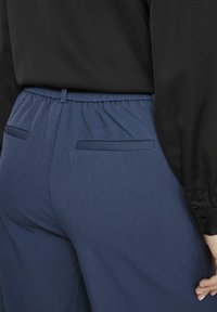 Modré kalhoty s elastickým pasem a dvěma zadními kapsami, v kombinaci s černou blůzou se krajkovými detaily na rukávech.