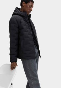 Giacca a vento nera con design trapuntato, chiusura con zip e cappuccio. Indossata con jeans grigi, appoggiata contro una sedia bianca.