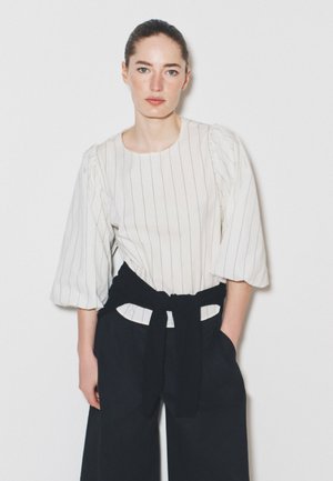 Femme portant une blouse blanche à fines rayures avec des manches bouffantes et un pantalon noir taille haute noué avec une ceinture noire, debout contre un mur blanc.