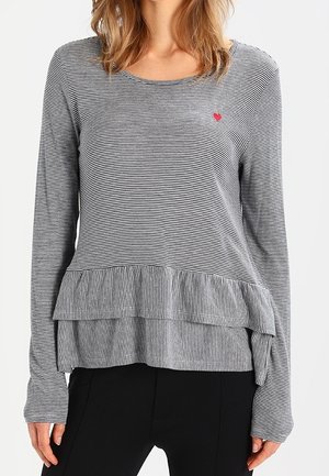 Kvinde iført en langærmet, sort og hvid tyndstribet top med lagdelte flæser ved kanten og en lille rød hjerte detalje på brystet.