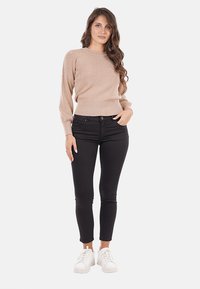 Donna in piedi con lunghi capelli mossi, indossa un maglione beige, pantaloni skinny neri e sneakers bianche, con una mano in tasca.