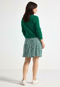 Grønt kabelstrikket sweater, blomstret nederdel med plisseringer, hvide sneakers. Outfitet har en tekstureret stof og livlige farver.