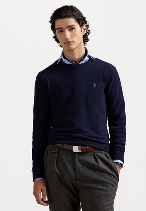 Marineblauwe wollen trui met ronde hals, voorzien van een klein logo op de borst; gedragen over een lichtblauw overhemd met kraag en grijze broek.