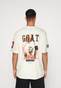 Koszulka w kolorze kremowym z dużym nadrukiem graficznym przedstawiającym zawodnika koszykówki, tekst "G.O.A.T." oraz szczegóły w czerwonym kolorze na rękawie.