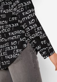 Chemise noire avec des motifs de texte blanc comportant des phrases telles que "MODE" et "VIE" dans des tailles variées. Manches longues, poignets à boutons et ourlet arrondi.