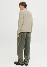 Pull en tricot beige avec un col haut, associé à un pantalon vert olive ample. Le mannequin se tient de dos, mettant en avant la coupe décontractée et le tissu texturé.