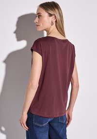 Burgundská krátká povlečená halenka s uvolněným střihem, kulatým výstřihem a bočními rozparky, v kombinaci s modrými džínami, zobrazeno zezadu.