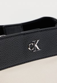 Μαύρη υφή δερμάτινης επένδυσης με ασημένιο λογότυπο "CK" στην κορυφή μιας μπεζ υφασμάτινης τσάντας, που δείχνει το άνοιγμα της τσάντας.