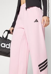 Lys rosa joggebukser med elastisk midje, med svart Adidas-logo og tre svarte striper på underbenet.