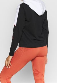 Svart sweatshirt med vit huva och röda detaljer; ihop med figurnära korallfärgad leggings med en strukturerad mönster. Nærbild bakifrån.