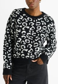 Schwarzer Pullover mit einem flauschigen weißen Leopardenmuster, Rundhalsausschnitt und gerippten Bündchen, kombiniert mit dunklen Jeans.