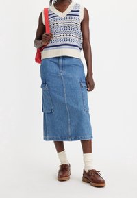 Pletená bezrukávová svetr s modro-krémovým vzorem, spárovaný s denimovou midi sukni s bočními kapsami a hnědými šněrovacími botami.