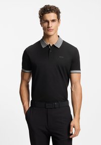 Czarna koszulka polo z wzorzystym szarym kołnierzykiem i mankietami, z zapięciem na guziki i logo na lewej piersi. Noszona z czarnymi spodniami i paskiem.