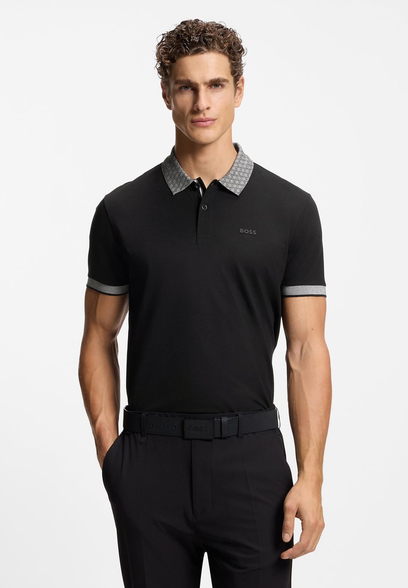 Czarna koszulka polo z wzorzystym szarym kołnierzykiem i mankietami, z zapięciem na guziki i logo na lewej piersi. Noszona z czarnymi spodniami i paskiem.