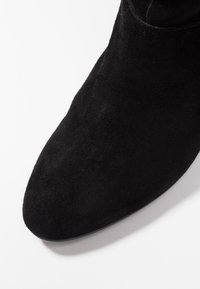 Botín de ante negro con punta afilada, exhibido sobre un fondo blanco, mostrando detalles de textura y forma.