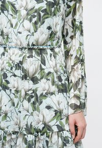 Jurk met bloemenpatroon van witte en groene magnolia's, doorschijnende lange mouwen en een dunne zilveren kettingriem met een hangende bedel.