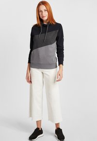 Felpa con cappuccio in navy, grigio e grigio chiaro; presenta un design a blocchi di colore diagonali, polsini a coste e dettagli con coulisse. Indossata con pantaloni bianchi.